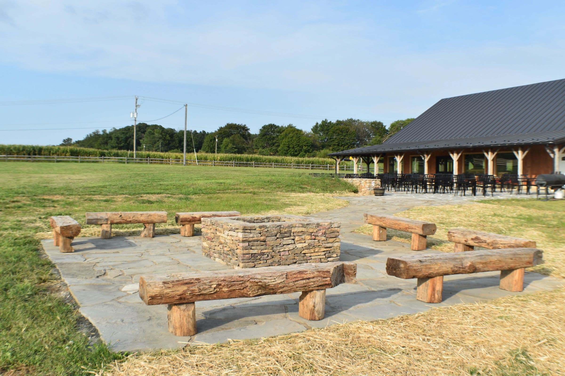 A stone fire pit ringed by log benches on the flagstone patio, with the timber-frame taproom and the fields beyond.
